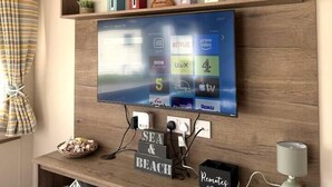 A living room with a mounted TV displaying streaming services, a wooden shelf with decorative items, and a remote control on the counter.