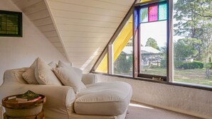 A room with a white ceiling, a large window with yellow curtains, a white sofa, and a small wooden table with books. - Petana Farm Stay - Pet-friendly Country Retreat (Morton)