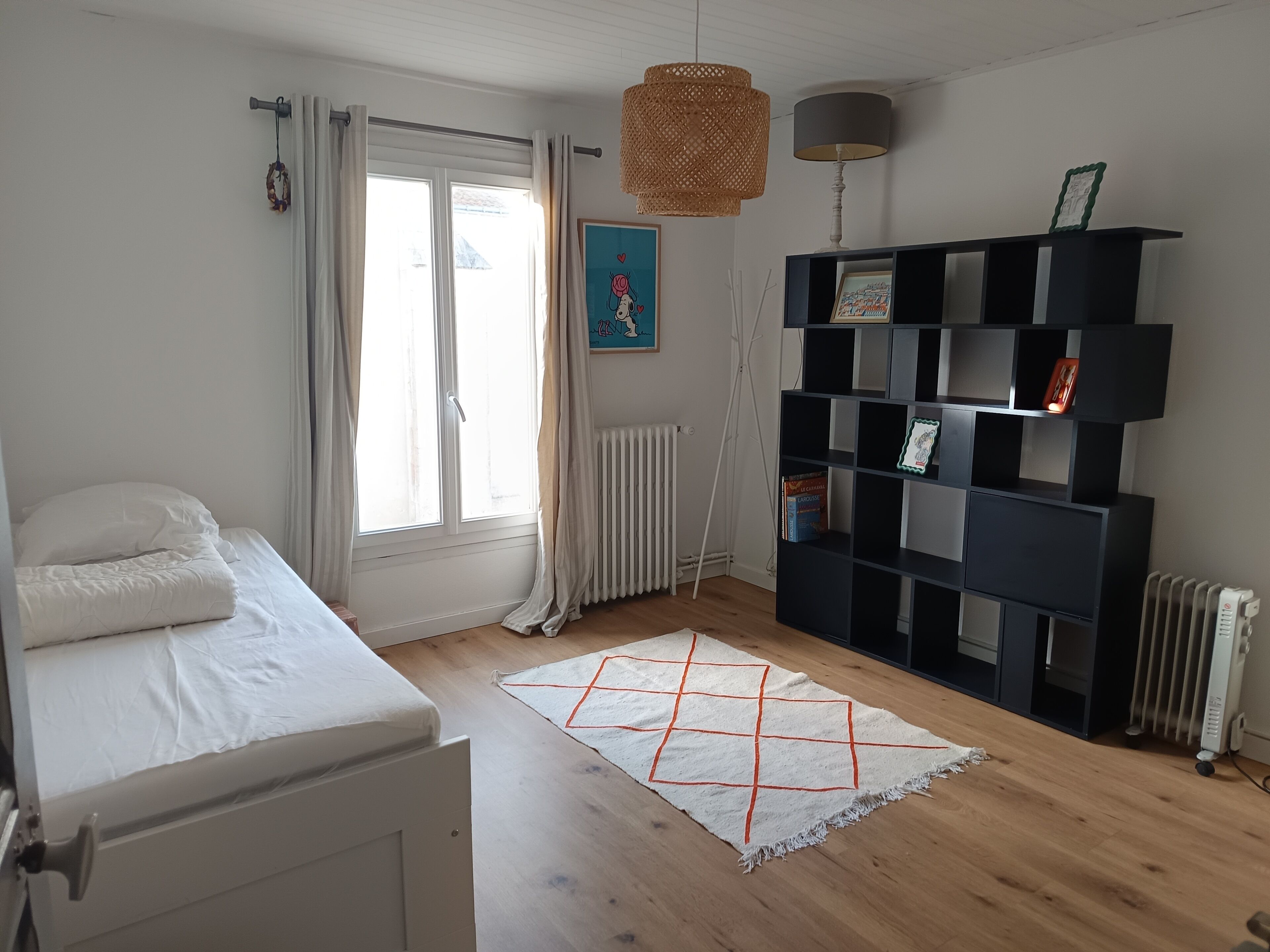 A bedroom with a bed, a window with curtains, a radiator, a bookshelf, and a hanging light fixture.
