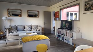 Un salon comprenant un canapé, un pouf, un téléviseur, une bibliothèque et une fenêtre donnant sur un beau paysage.