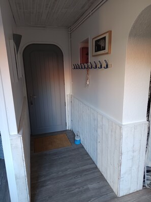 Een gang met een houten deur, een blauwe emmer en een kapstok met haken.
