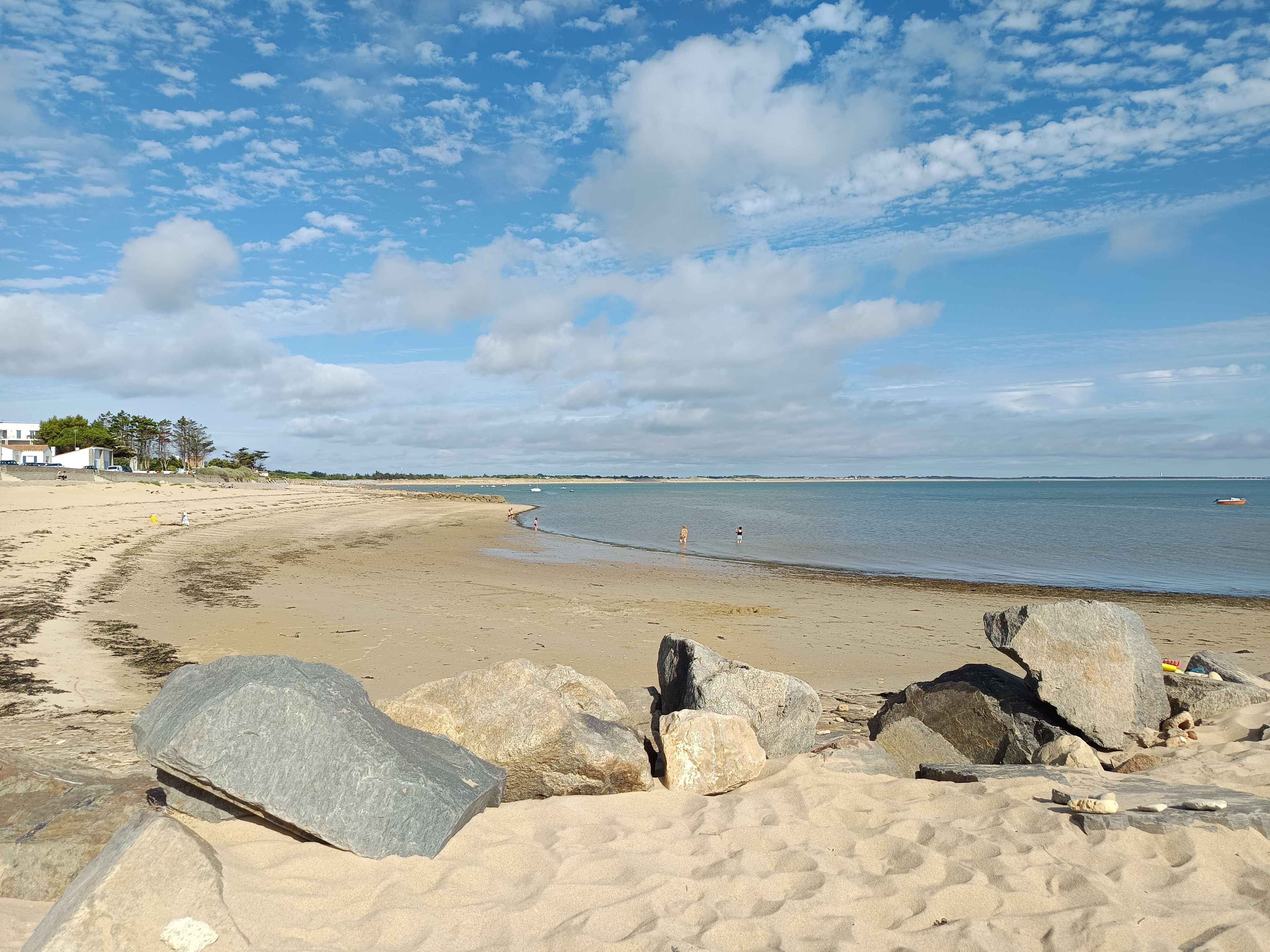 A beach with large rocks in the foreground, a sandy shore, and a calm sea under a partly cloudy sky.
