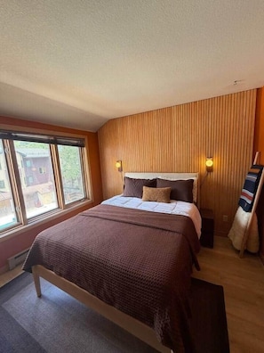 A bedroom with a large bed, a window, a wall-mounted light, and a wooden panel wall. - The Western View: Pool, Hot Tub, Ski, Repeat (Telluride)