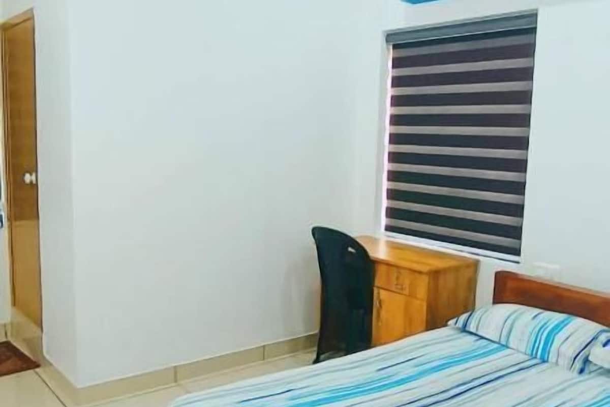 Un dormitorio con cama, escritorio, silla, ventilador de techo y ventana con persianas.