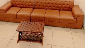 Un sofá marrón con respaldo capitonado y una mesa de centro de madera en una habitación con paredes blancas y suelo de baldosas.