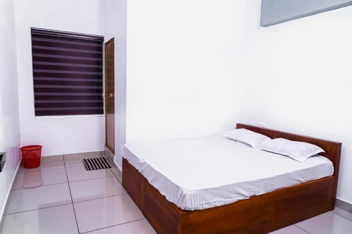 Un dormitorio con cama de madera, ropa de cama blanca, una ventana con persianas, una cesta roja y suelo de baldosas.
