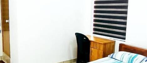 A bedroom with a bed, a desk, a chair, a ceiling fan, and a window with blinds.