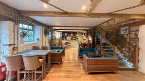 Un interior rústico con vigas de madera a la vista, una zona de estar central con sofás y una mesa, y una escalera que conduce a un nivel superior.