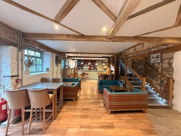 Ein rustikaler Innenraum mit freiliegenden Holzbalken, einer zentralen Sitzgruppe mit Sofas und einem Tisch sowie einer Treppe, die in einen oberen Bereich führt.