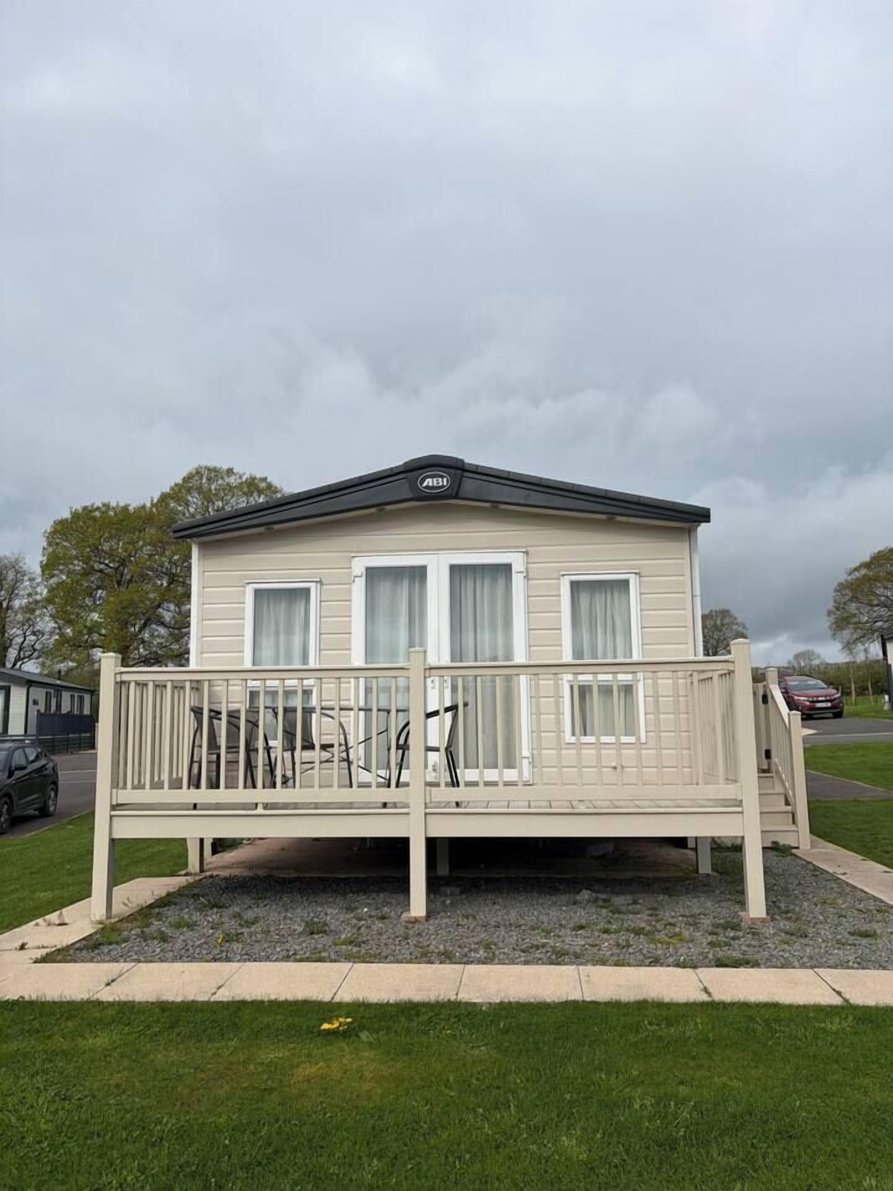 A single-story mobile home with a front deck, a covered parking area, and a grassy lawn.