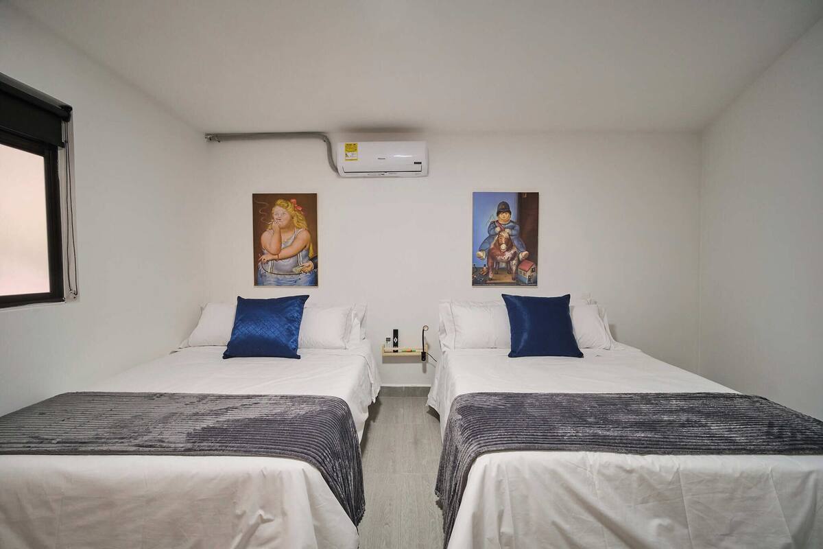 Quarto de hotel com duas camas, roupa de cama branca, almofadas azuis, uma mesinha com abajur e dois aparelhos de ar condicionado fixados na parede.