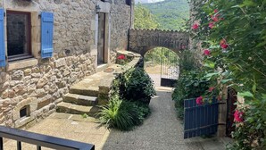 Un bâtiment en pierre doté d’une entrée étroite, d’une petite fenêtre avec des volets bleus et d’une arche en pierre menant à un jardin.