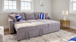 A grey sectional sofa with a furry rug, a small round table with a lamp, and a framed picture on the wall. - Luxury Beach House, Rooftop Deck, Ocean Views, Walk to Beach & Boardwalk (Seaside Heights)