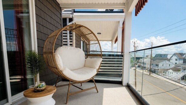 A balcony with a wicker chair, a small round table with a plant, and a view of houses through a glass railing. - Luxury Beach House, Rooftop Deck, Ocean Views, Walk to Beach & Boardwalk (Seaside Heights)