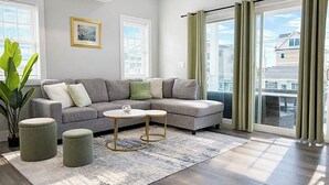 A modern living room with a grey sectional sofa, two round tables, a large window with curtains, and a potted plant. - Luxury Beach House, Rooftop Deck, Ocean Views, Walk to Beach & Boardwalk (Seaside Heights)
