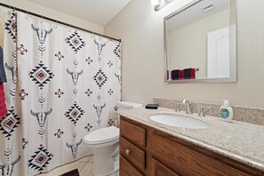A bathroom with a shower curtain featuring a geometric pattern, a toilet, a sink with a soap dispenser, and a mirror above the sink. - Family Retreat with Pool and Game Room (Avondale)
