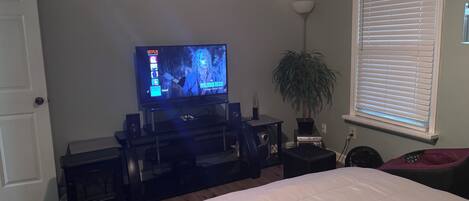 A bedroom with a bed, a TV on a stand, a plant, and a window with blinds.
