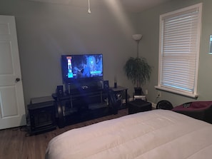 A bedroom with a bed, a TV on a stand, a plant, and a window with blinds.