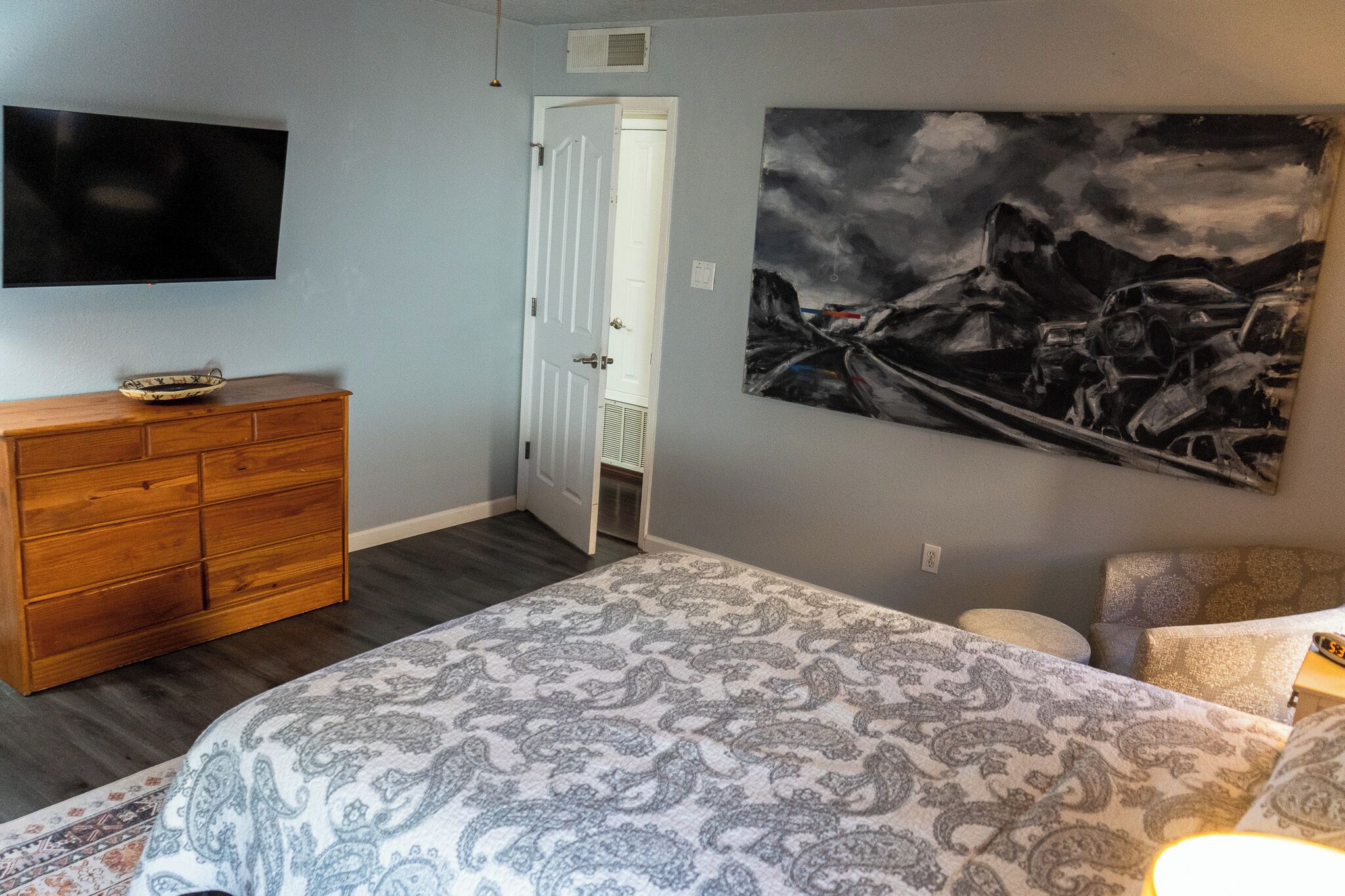 A bedroom with a bed, dresser, painting, and a door leading to another room.