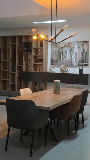 A modern dining area with a long table, chairs, a pendant light, and a bookshelf with various items. - Luxurious Contemporary Villa With Private Pool in Tunis (La Soukra)