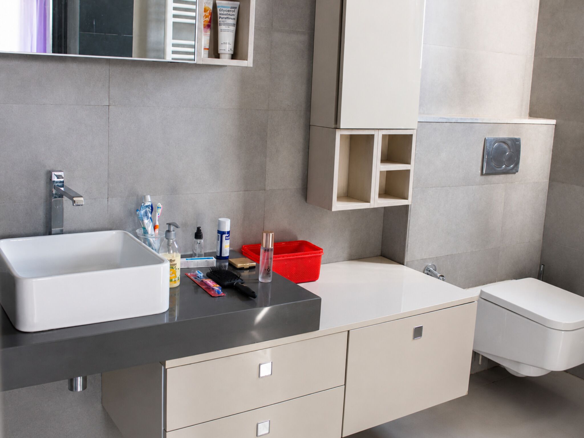 Un baño moderno con un lavamanos grande, un inodoro y un espejo. Hay artículos de aseo en la encimera y una cesta roja.