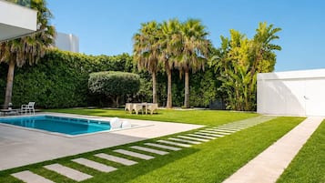 A modern outdoor pool area with a clear sky, neatly trimmed grass, and palm trees.