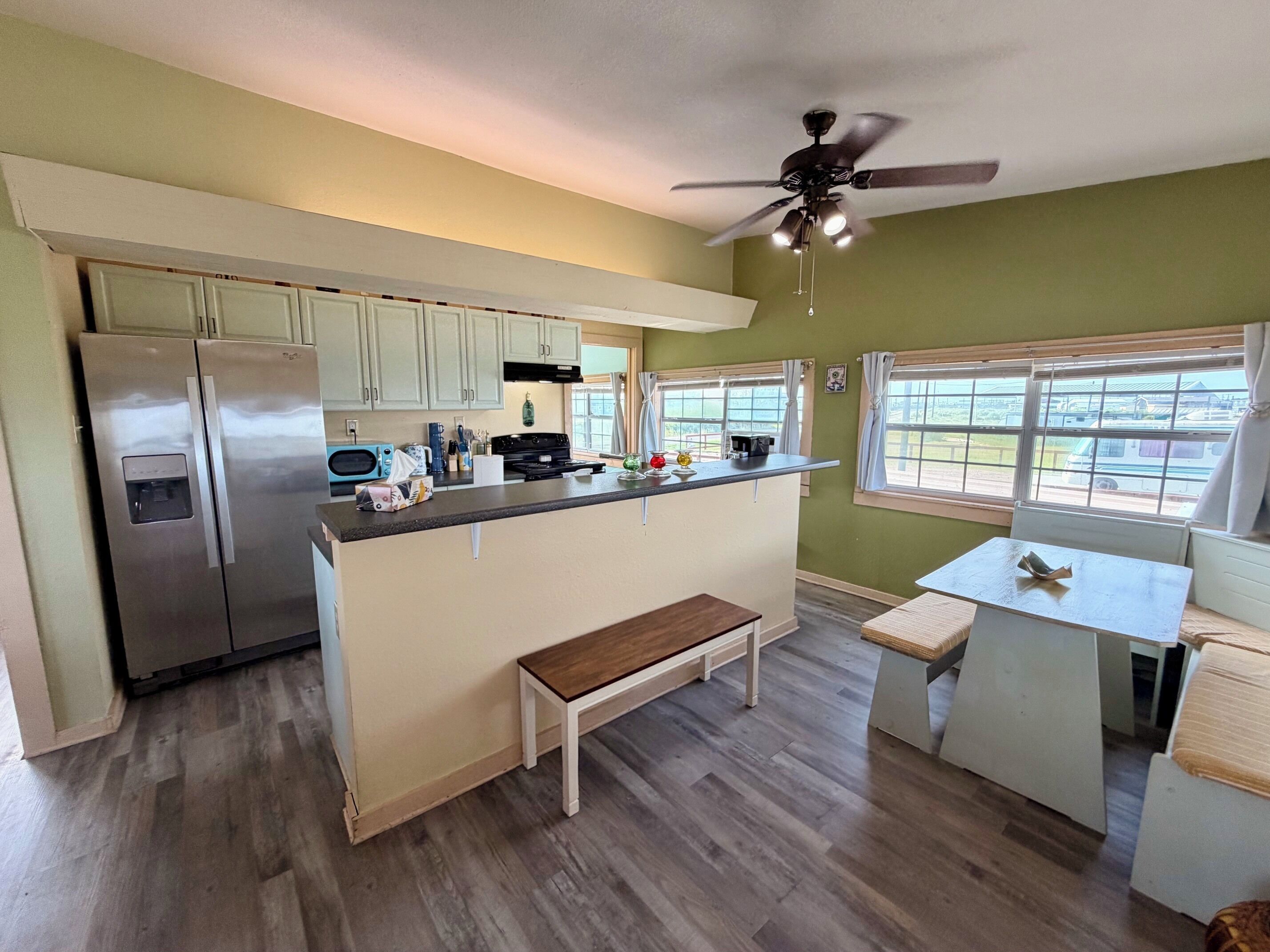 A kitchen with a refrigerator, microwave, and stove. There is a window with curtains, a ceiling fan, and a bench.