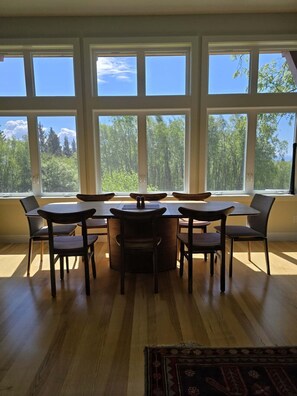 Ett matrum med ett träbord och stolar, stora fönster med utsikt över skogen och en mönstrad matta på golvet.