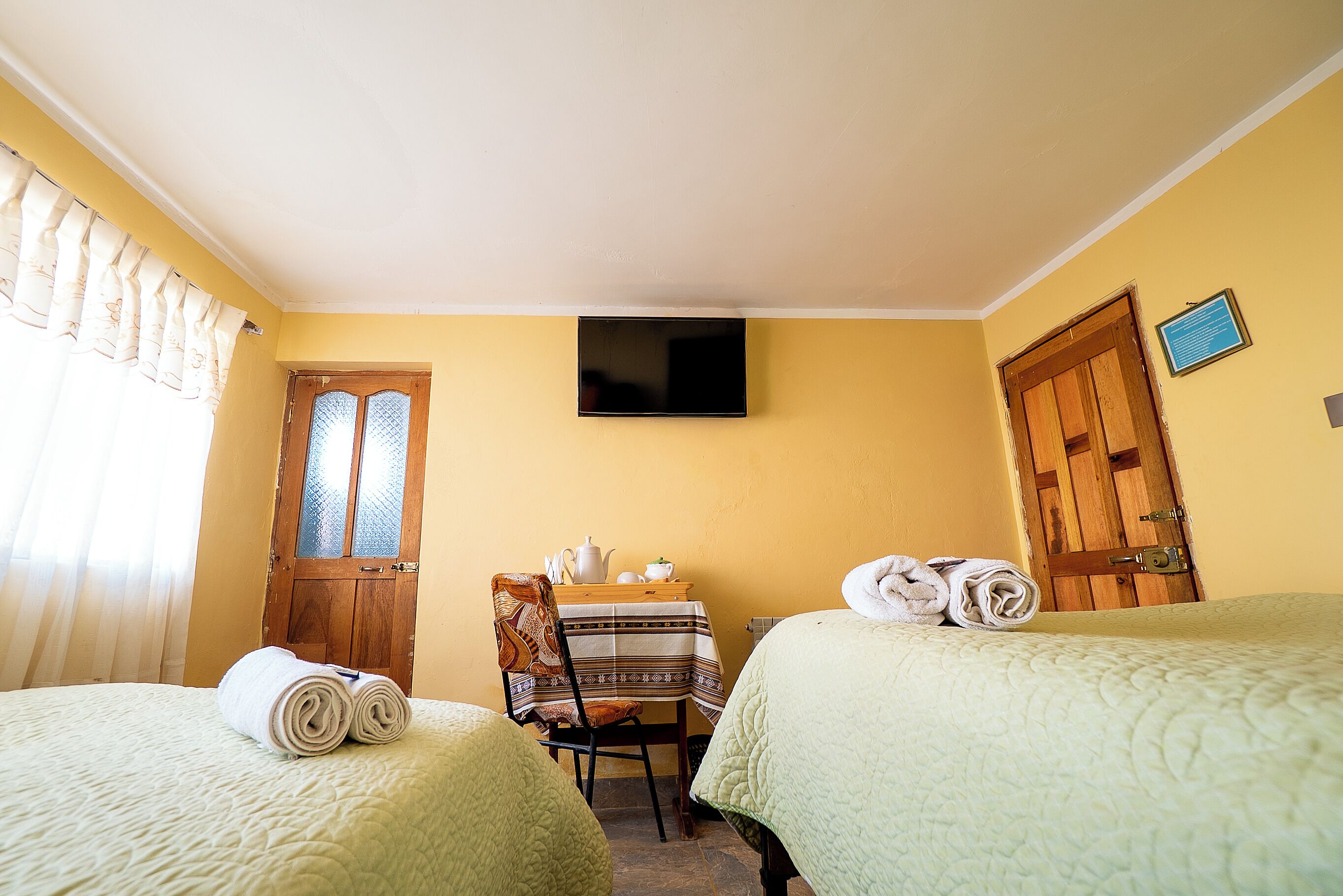 Une chambre avec deux lits, une table avec une chaise, un téléviseur fixé au mur et une fenêtre avec des rideaux.