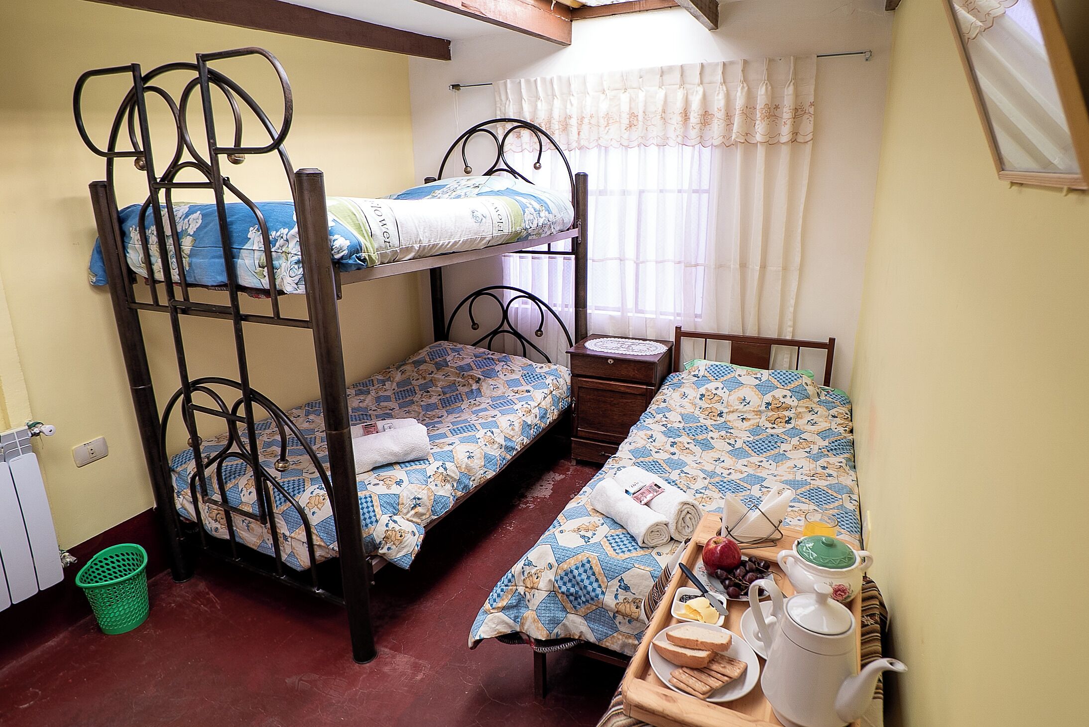 A dormitory room with bunk beds, a small bedside table with a kettle, a tray with breakfast items, and a window with curtains.