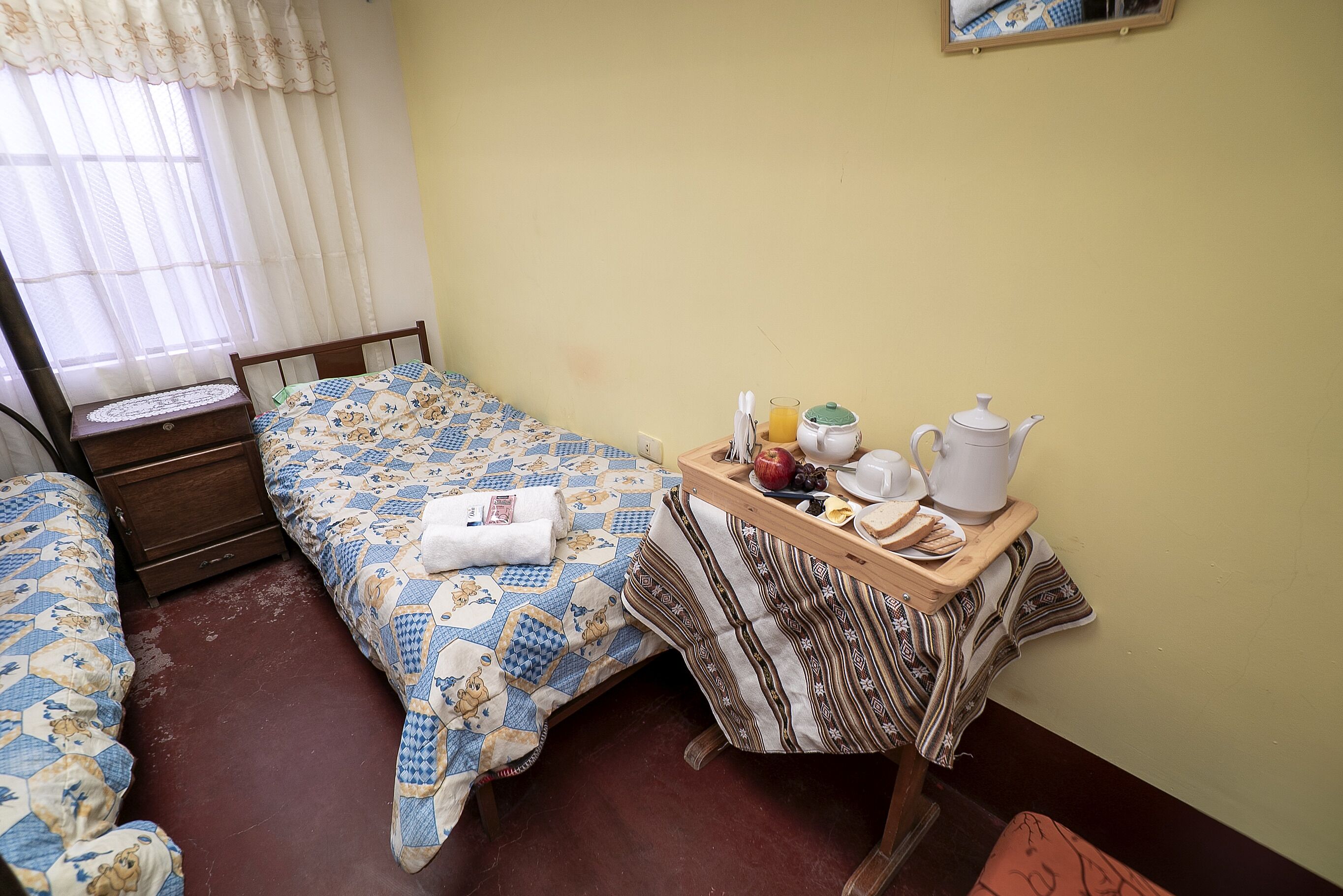 A bedroom with a bed, a bedside table with a tray of breakfast items, and a window with curtains.