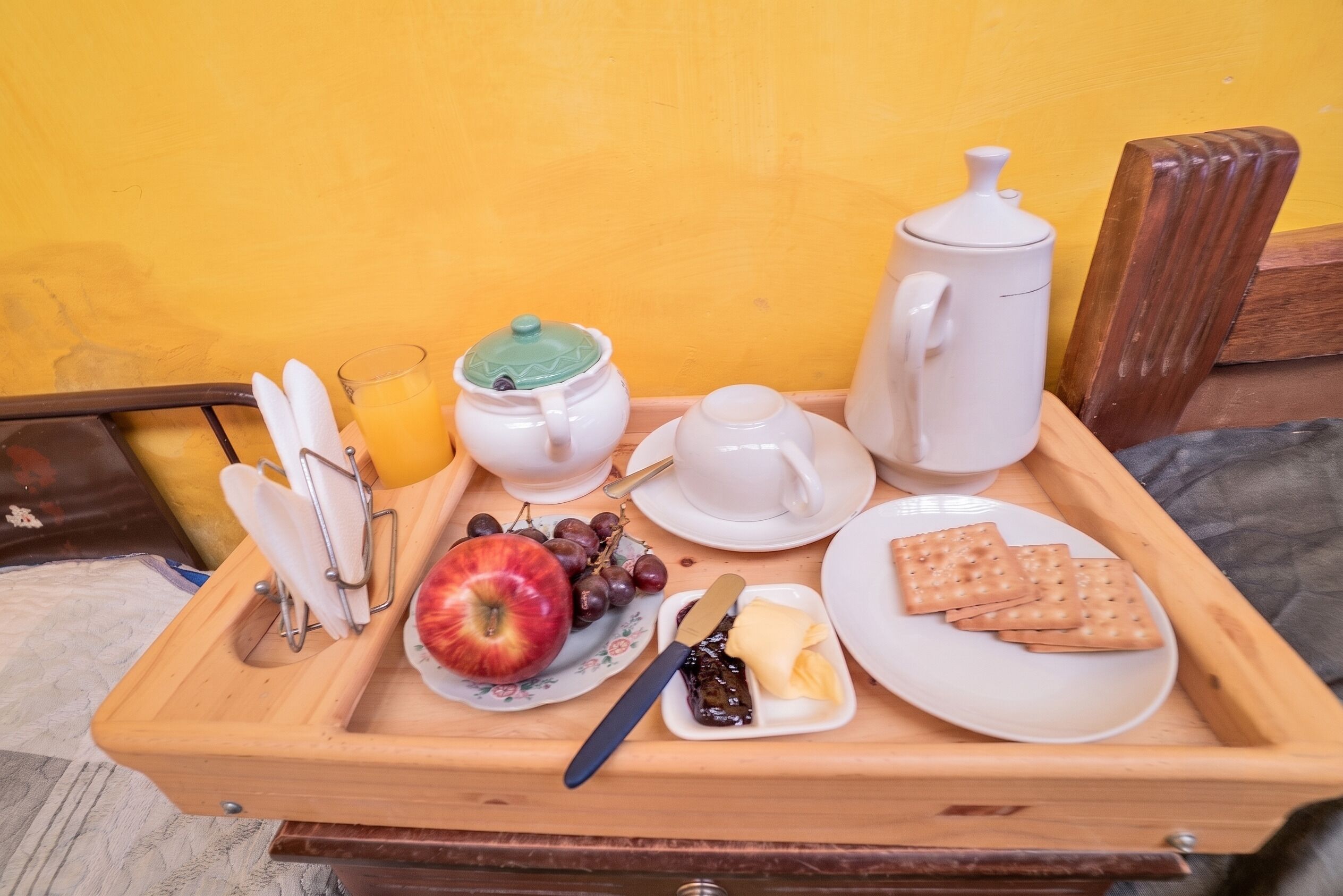 A wooden tray with breakfast items including a teapot, cups, a jug, a plate with an apple, grapes, butter, jam, and biscuits.