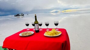 Una mesa con una botella de vino, copas y platos de comida sobre un mantel rojo, con un telón de fondo de playa y un barco a lo lejos.