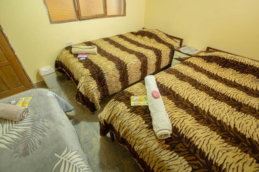 A room with two beds, each with a patterned bedspread, a small table with a lamp, and a window with blinds.
