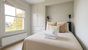 Une chambre avec une grande fenêtre, un lit avec une literie beige, deux lampes de chevet et un radiateur.