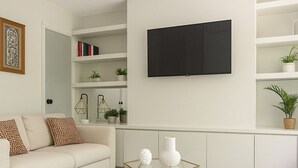 Un salon comprenant un canapé, une table basse, un téléviseur, des étagères avec des livres et des plantes, et un tapis à motifs.