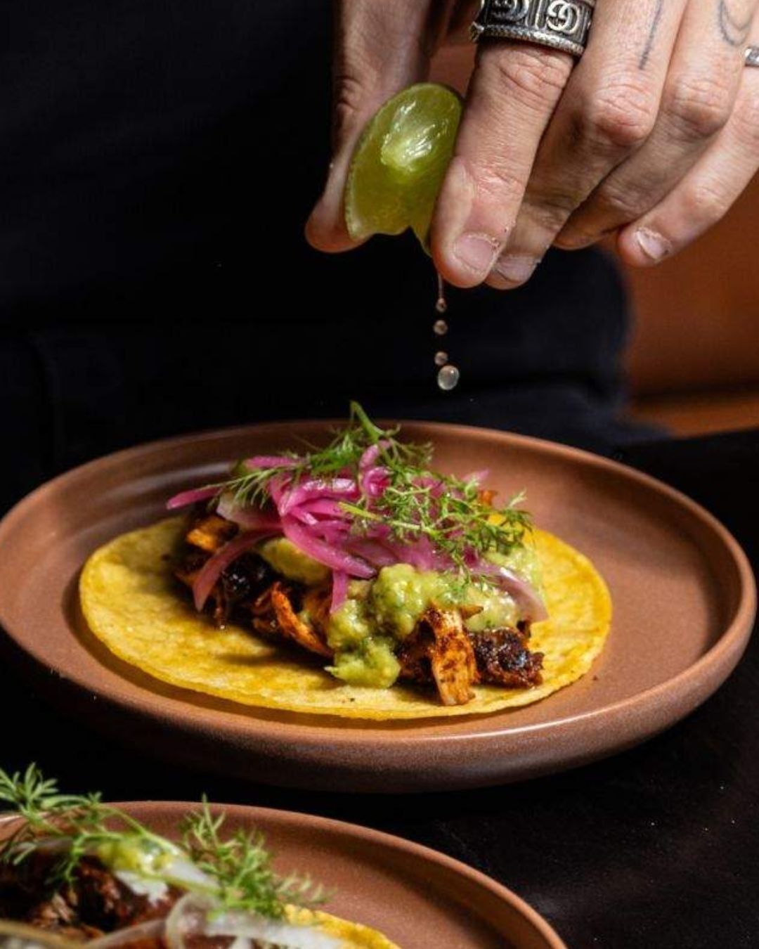 Eine Person gibt eine grüne Soße auf ein Gericht mit Tortilla-Basis, belegt mit Fleisch und garniert mit roten Zwiebeln und Kräutern.