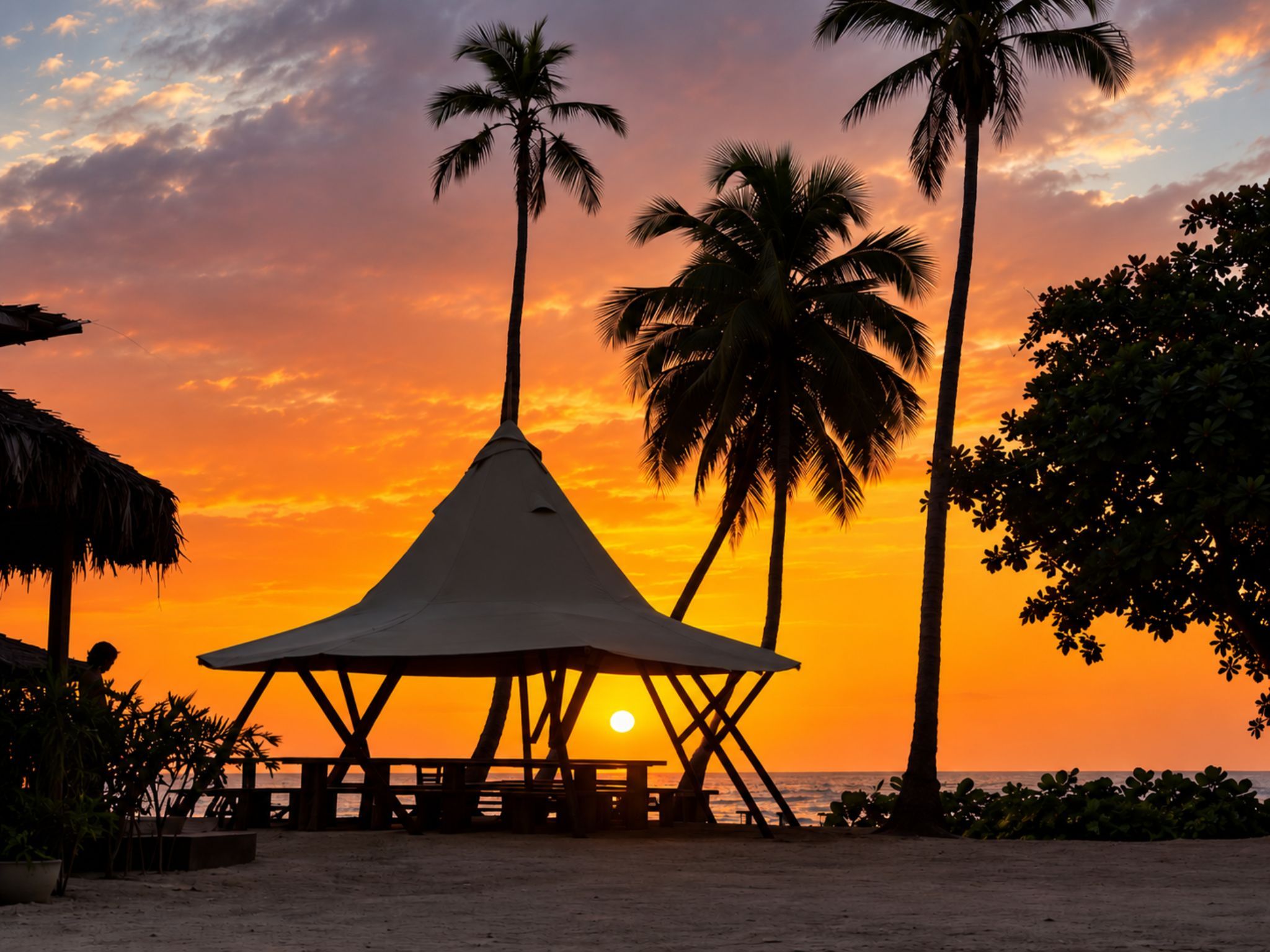 A beach scene with a thatched-roof structure and palm trees at sunset.