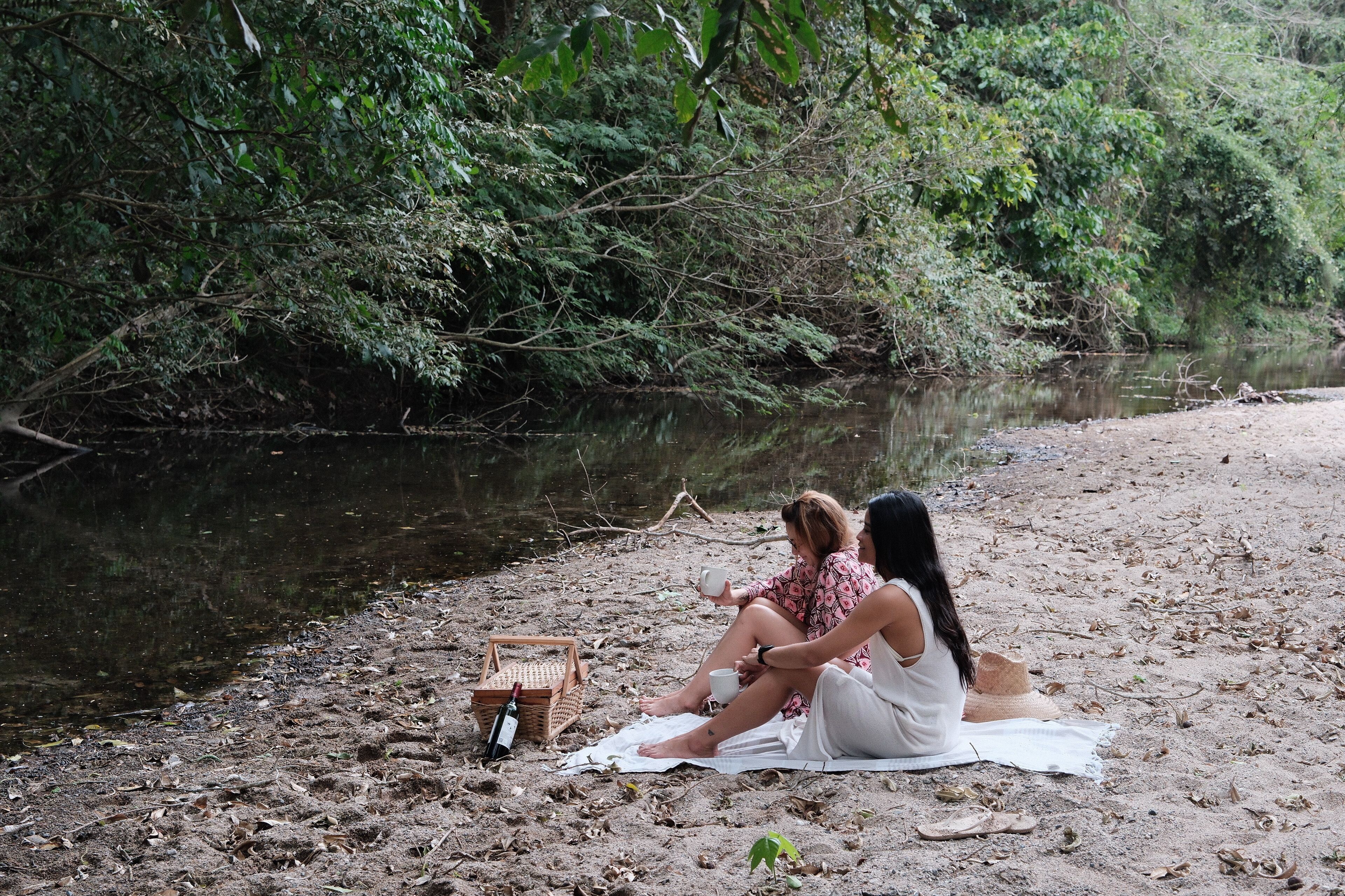 Dos personas sentadas sobre una manta junto a un río, con una cesta de picnic y una botella cerca.
