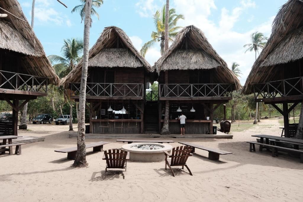 A beachside resort with thatched-roof huts, outdoor seating, and a fire pit.