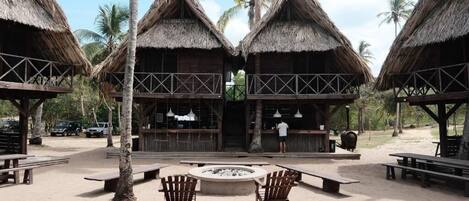 A beachside resort with thatched-roof huts, outdoor seating, and a fire pit.