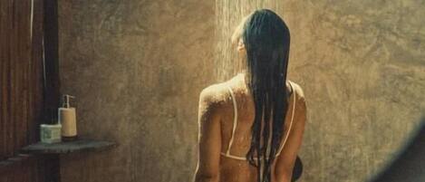 Una persona en una bañera con ducha sobre ella, un estante de madera con objetos y una pared con textura.