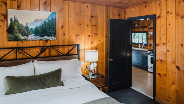 A wooden-paneled room with a bed, a nightstand with a lamp, a painting of a forest, and an open door to a kitchen. - 2 BR 2 BA San Juan Mountain Cabin (Archuleta County)