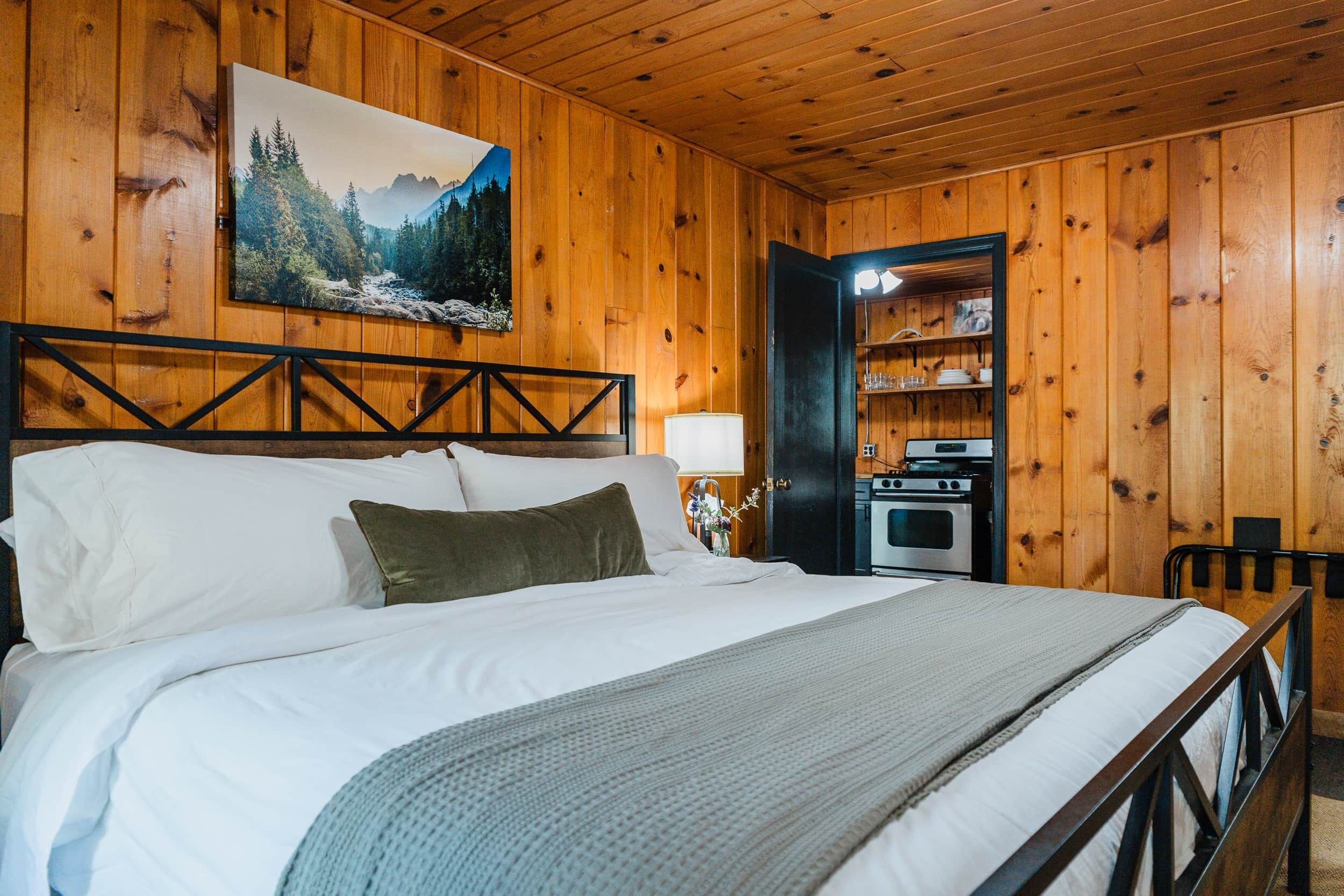 A wooden-paneled room with a bed, a painting of a forest, and a kitchen area.