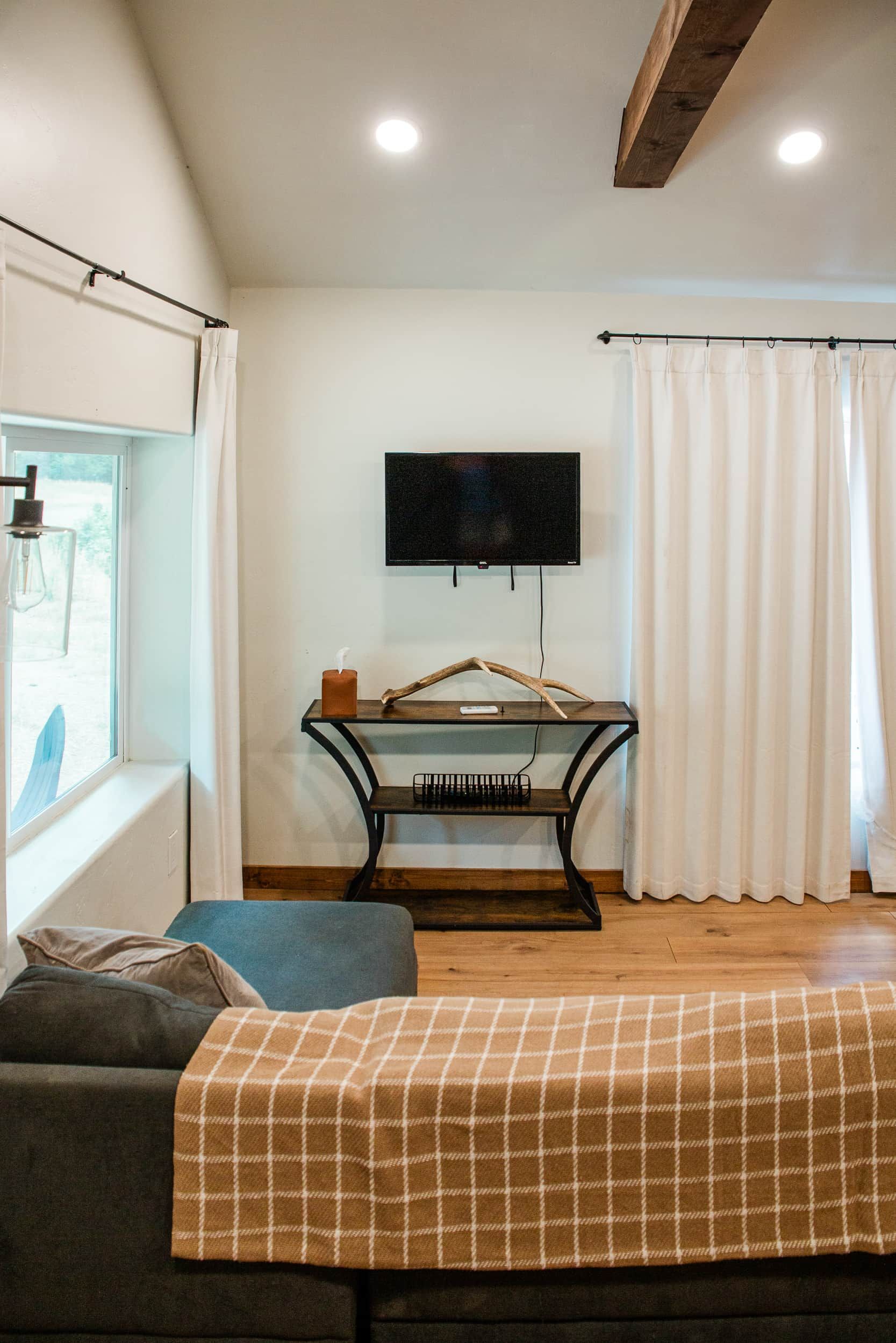 A room with a flat-screen TV mounted on the wall, a wooden table with a candle and a remote control, a window with curtains, and a sofa with a plaid blanket.