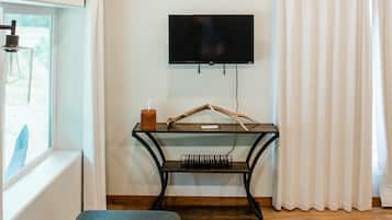 A room with a flat-screen TV mounted on the wall, a wooden table with a candle and a remote control, a window with curtains, and a sofa with a plaid blanket.