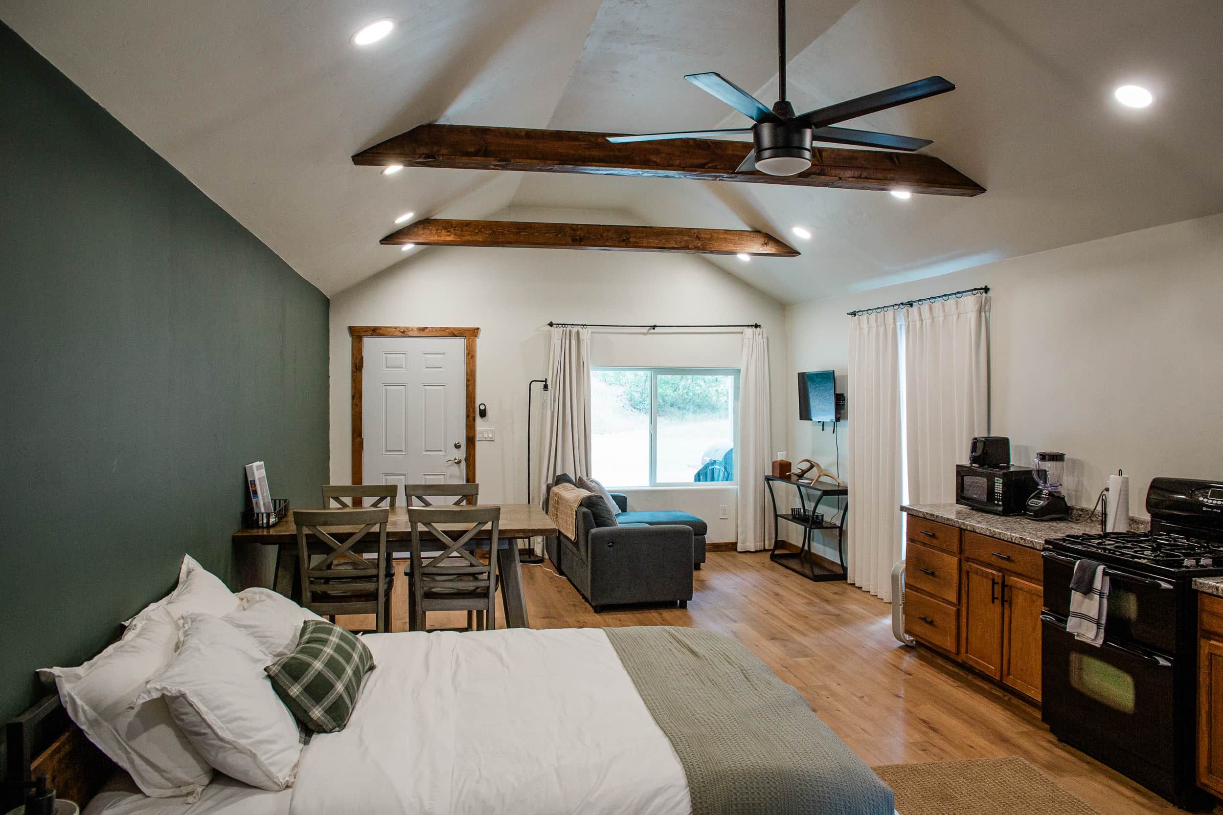 A compact room with a bed, a dining table, a kitchenette, and a ceiling fan.