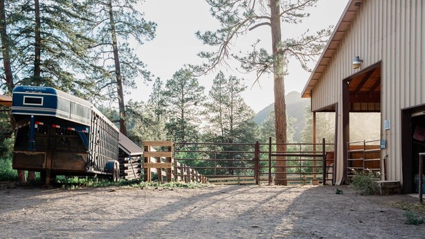 A dirt road leading to a wooden building with a covered area, surrounded by trees and a blue vehicle.
