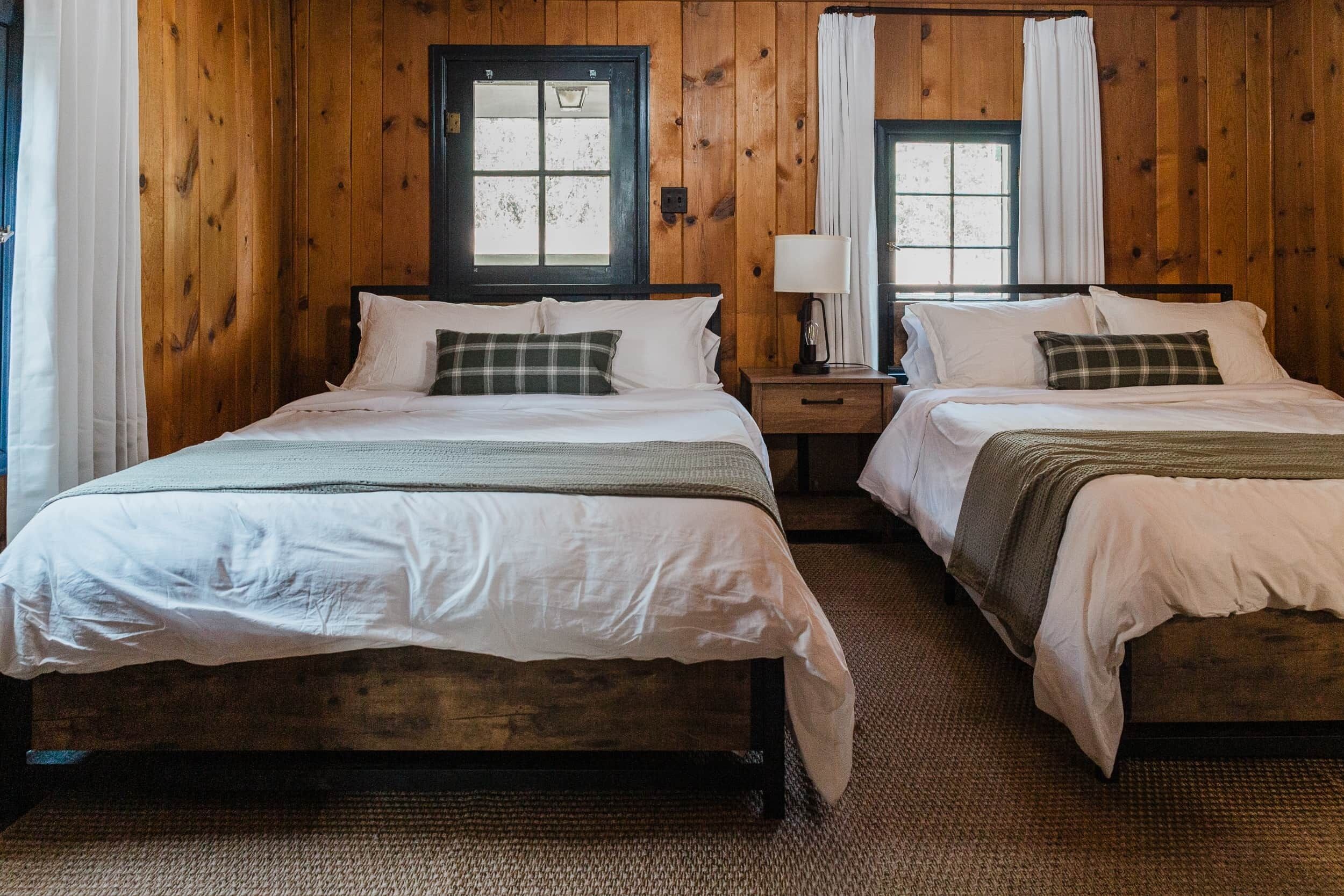 A room with two wooden beds, a nightstand with a lamp, and a window with curtains.