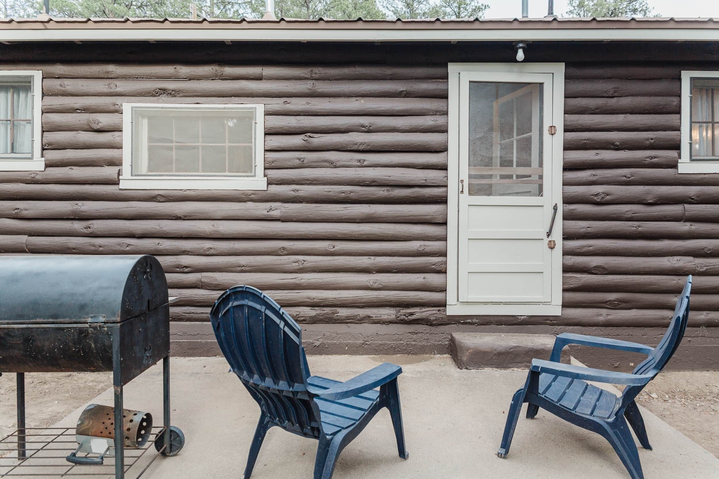 A log cabin with two blue plastic chairs, a grill, and a door.